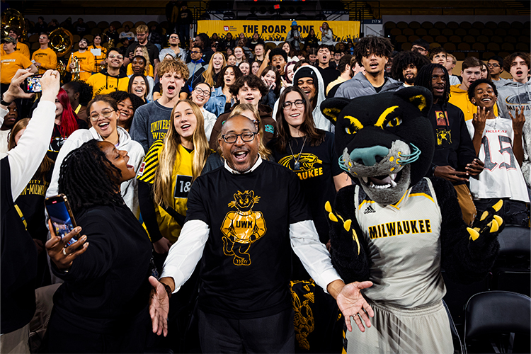 Chancellor Gibson stands with students cheering in the stands at a basketball game.