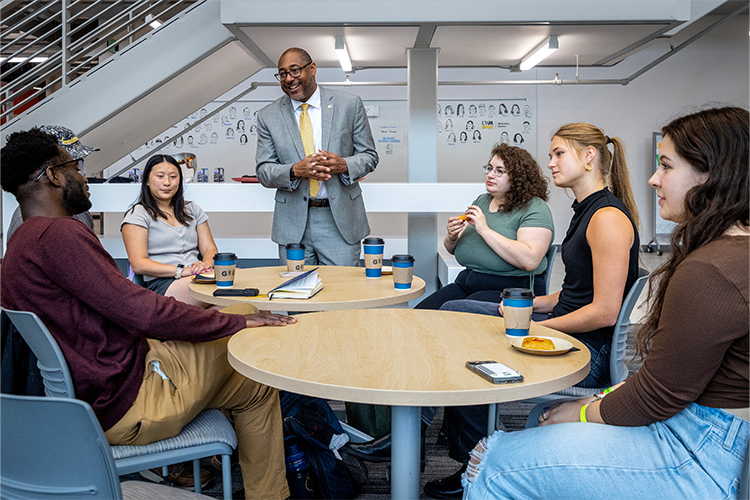 Chancellor Gibson talks with students who are sitting around tables.