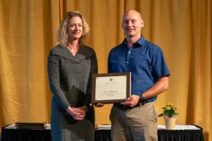 A woman and a man stand in front of a curtain. The man is accepting an award plaque