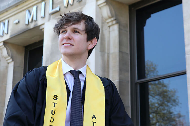 White male student wearing graduation gown and smiling into distance