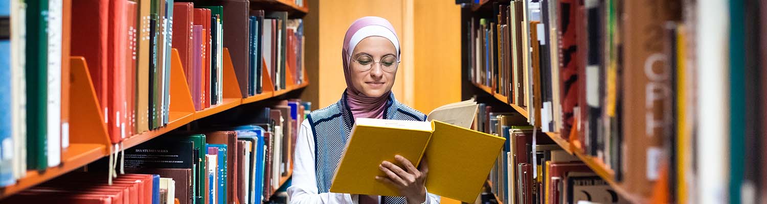 Female student in hijab looking at a book in a library