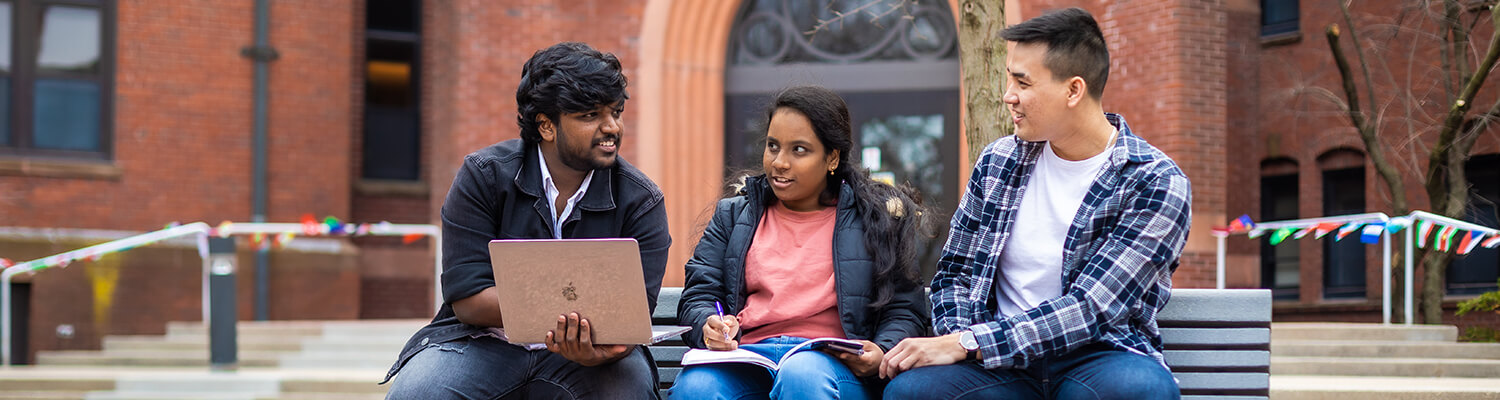 Three international students sitting on bench looking at laptops and talking to each other