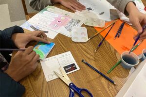 Close up of a table filled with art supplies, with two sets of hands working on making zines