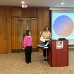 Two people standing at the podium speaking and smiling. The screen behind them is titled Dance Party!