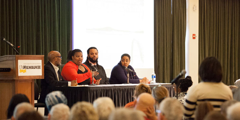 African-American professor and community members at a panel discussion with large audience.