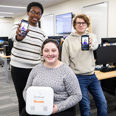 Group of Information Studies students (Black female, white male, white female) showing their phones in a tech lab environment.