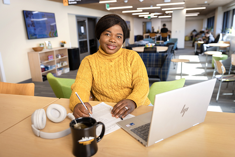 Female student sitting at table with laptop, headphones, notebook, and coffee. Student is smiling at the camera.