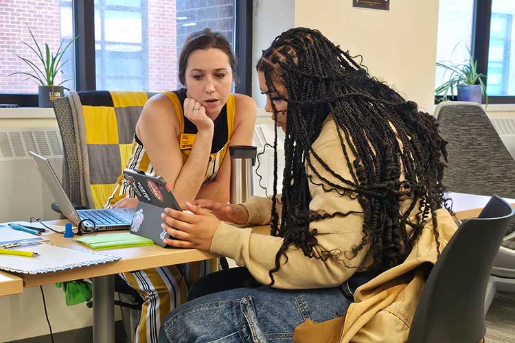 Female academic advisor and student lean in to review something on a laptop computer, collaborating at a table filled with notes, a laptop. A focused work session.
