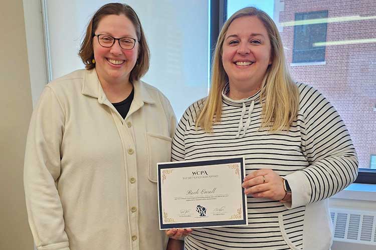 Two females standing in front of a window. One is holding a paper certificate. Both are smiling.