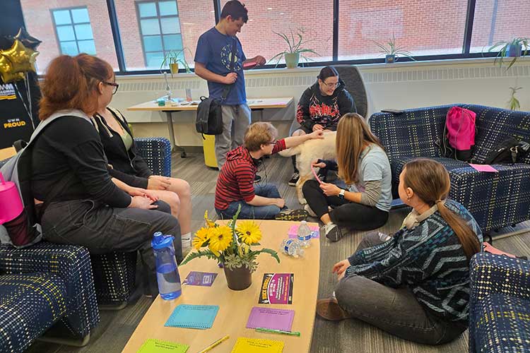 Group of students - some sitting on couches while other sit on the floor. Some are petting a yellow lab as others watch on.