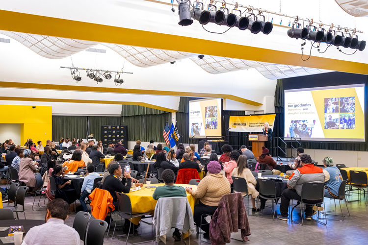Large room filled with attendees sitting at tables and listening to ceremony introduction by Dean Tina Freiburger, who is standing at a podium