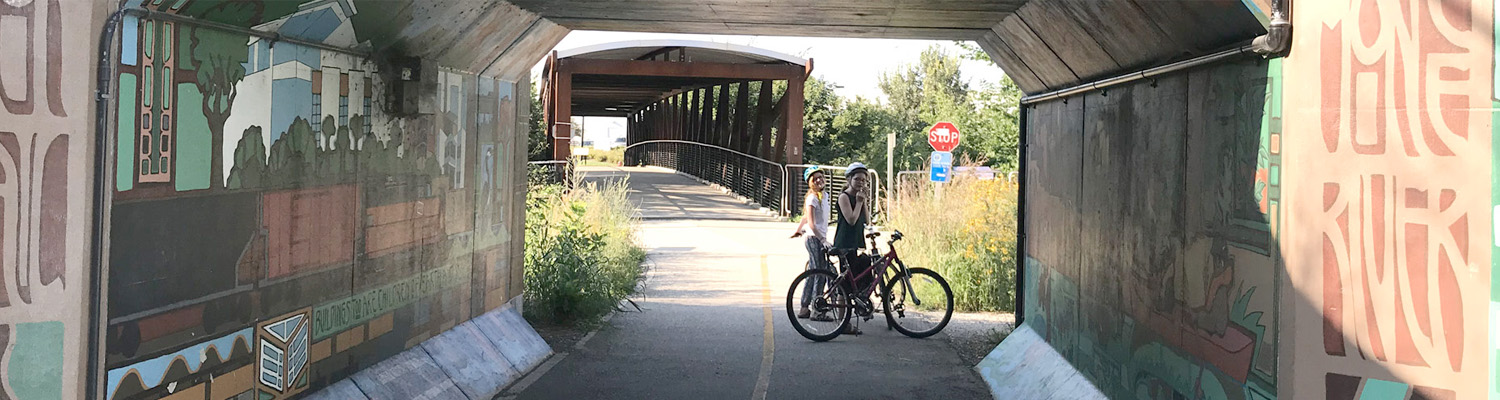 Two cyclists with bikes standing under a covered bridge on a paved trail with colorful murals on the concrete walls