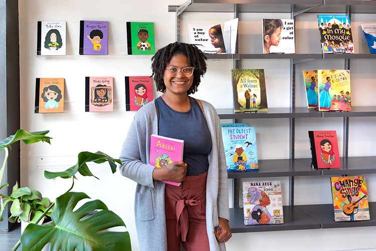 Ashley Valentine (african american woman) standing in front of a shelf of books at her Rooted MKE bookstore and tutoring center.