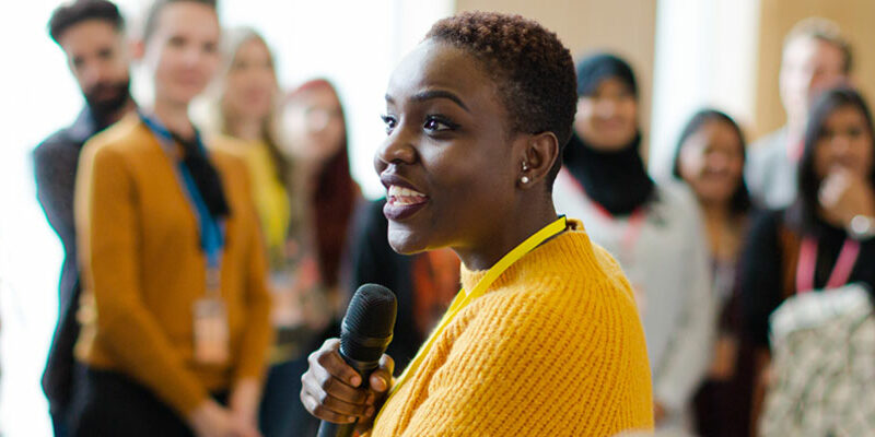 Smiling female speaker (black woman) with microphone in front of diverse audience.