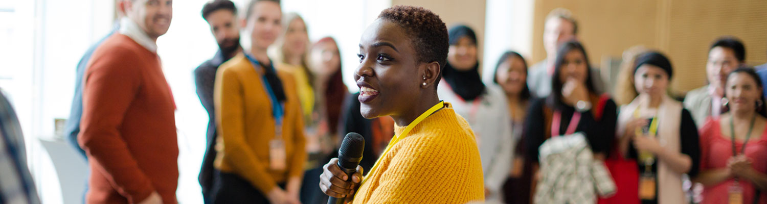 Smiling female speaker (black woman) with microphone in front of diverse audience.