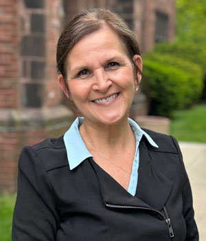 Professional headshot in natrual lighting of a professor (white female) standing outside an old brick campus building with green foliage all around.