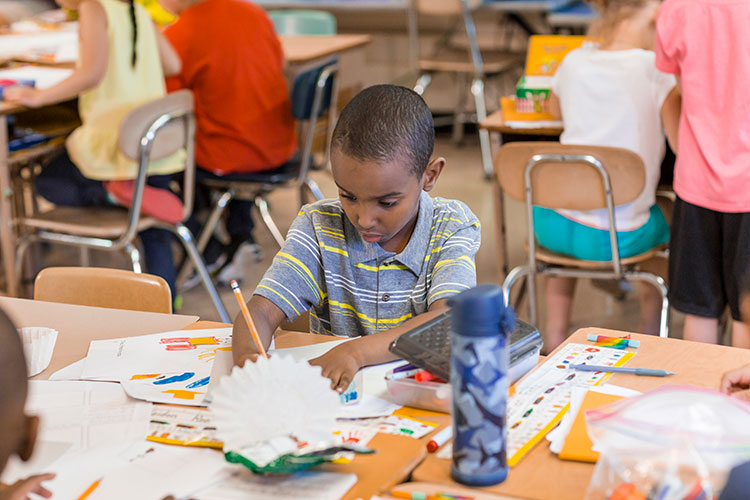 A young child (black male) working on a project at his desk in his classroom
