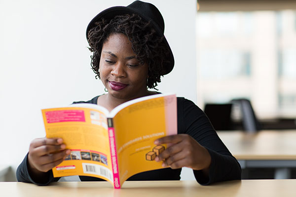 A black woman wearing a hat is reading a book while sitting at a table
