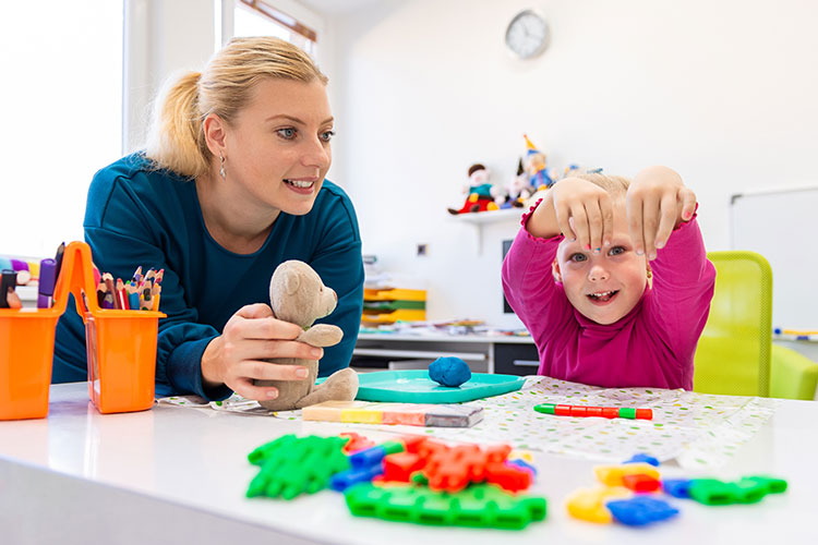 Teacher (white woman) observing a  happy child playing with items on her desk.