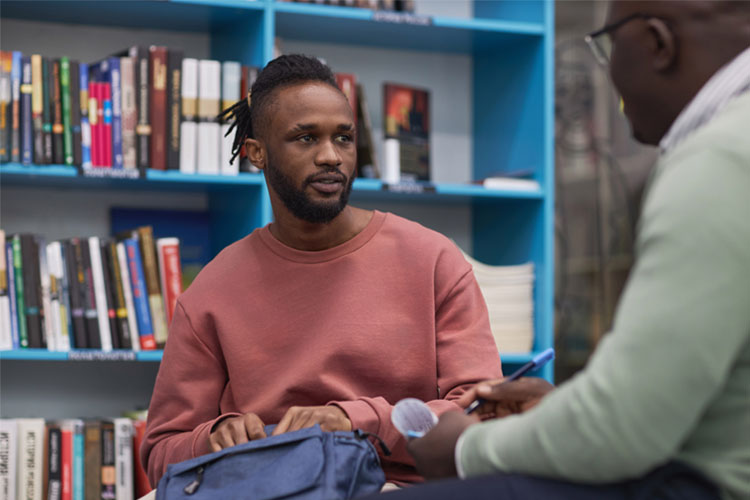 Portrait of black young man talking to advisor (black male) in college library.