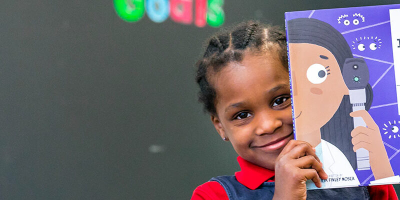 Young student (african american girl) holding a book up while smiling for the camera