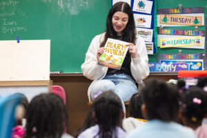 Teacher (white woman) sitting on a chair holding a book titled, "Diary of a Worm" while teaching early childhood literacy to a classroom of children.