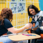 Two female university students (Black women) chatting in front of a gold wall map of Milwaukee.