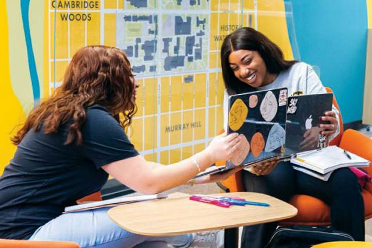 Two female university students (Black women) chatting in front of a gold wall map of Milwaukee.