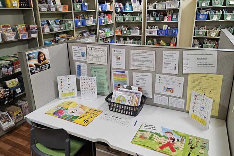 Room with shelves of books lining the walls. The center of the room contains cubicles set-up with two chairs designed for one-to-one tutoring sessions. The desk features brightly colored children's books.