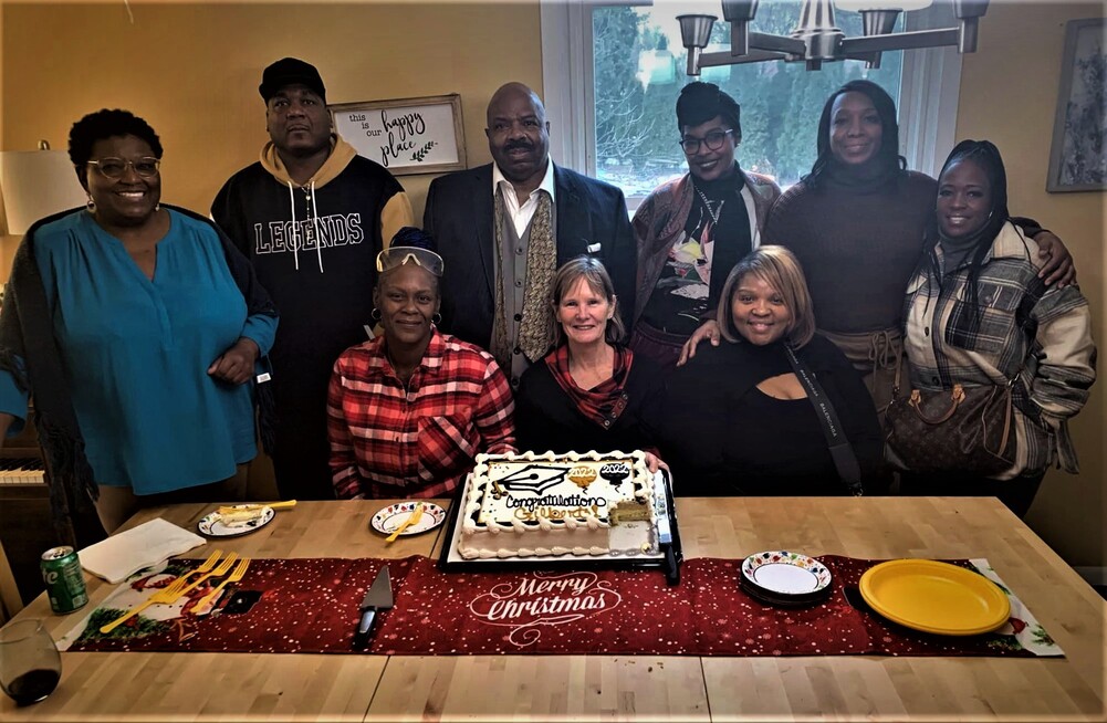 graduation party setting around a kitchen table with a cake in the center. A row of 6 individuals stands behind the table with three individuals seated at the table in front of them.