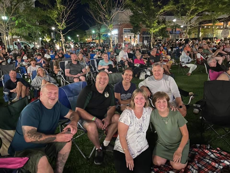 large outdoor gathering of people. focus of camera is on 6 individuals sitting in lawn chairs, some kneeling while smiling at the camera