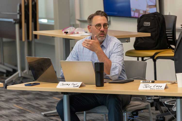 Man with glasses and a beard sits at a desk with a laptop and a tumbler, gesturing while speaking.