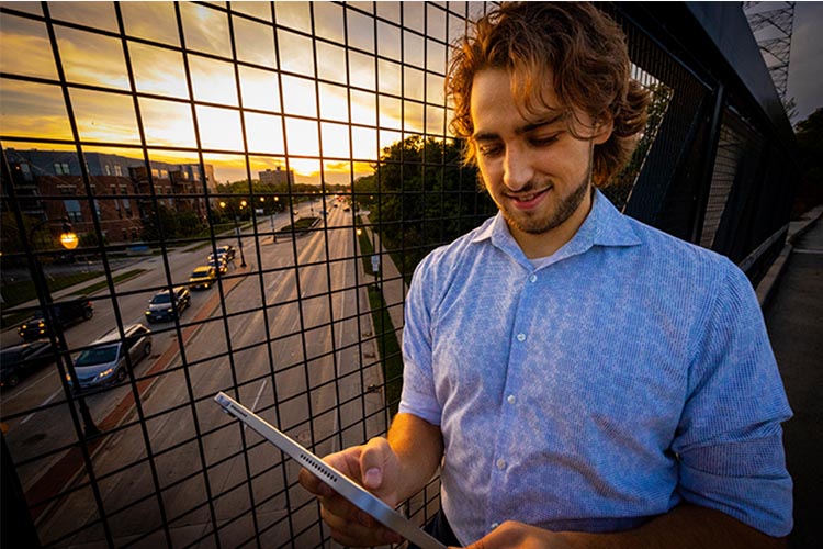 A man with shaggy brown hair and a light blue shirt looks at his tablet. A modern glass side of a building behind him reflect road traffic.
