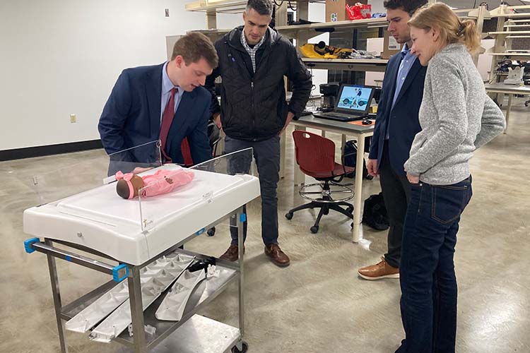 Three men and one woman look over an infant bed that that was modified. An infant doll lies on the bed. Three men and one woman look over an infant bed that that was modified. An infant doll lies on the bed.