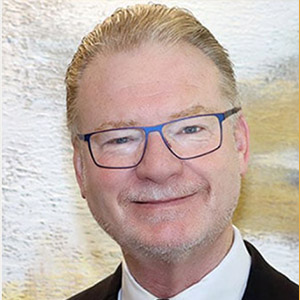 A headshot of a white older man with glasses and blonde-gray hair, slicked up.