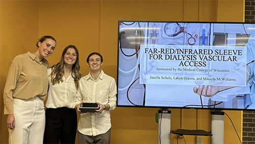 Three young women look at the camera. The one on the right is holding a small piece of equipment and stands next to an image on a screen.