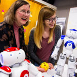 A female professor and a female student share small robots that can communicate. One robot is white and red and the other white and blue.