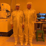 Two men in white suits pose in a orange-tinted clean room. Both are wearing glasses.