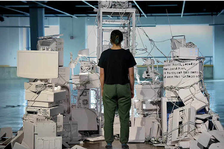 the back of a young woman in green pants who is standing in front of an art installation that looks like a pile of computer junk.