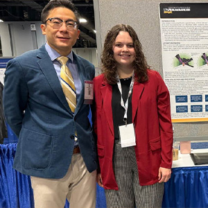 A man in a blue sports coat and a woman in a red sweater pose in front of a poster, both looking at the camera.