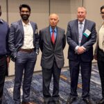 Five men standing in a line looking at the camera. Three are wearing dark sports coats, one a navy sweater and the fifth a light-colored jacket.