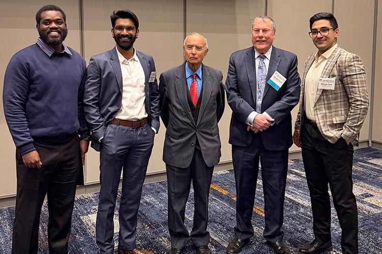 Five men standing in a line looking at the camera. Three are wearing dark sports coats, one a navy sweater and the fifth a light-colored jacket.