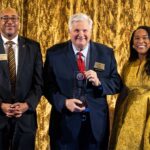 Two men, one with glasses, and one woman wearing a gold dress, display an award plaque while looking at the camera.