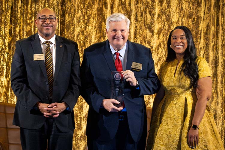 Two men, one with glasses, and one woman wearing a gold dress, display an award plaque while looking at the camera.