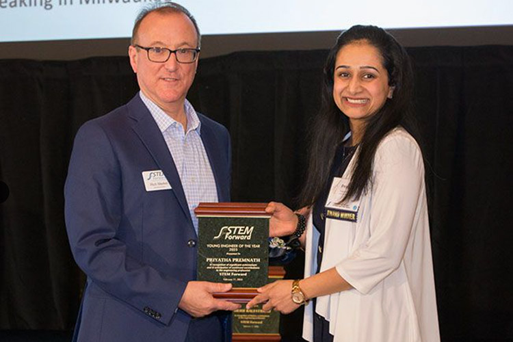 A man in a blue jacket and glasses presents a plaque to a woman with long dark hair and a white sweater. Both are looking at the camera.