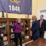 Three men in dark suit jackets and one women in a purple jacket talk among themselves.