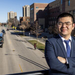 A man with glasses and a navy sportscoat and tie looks at the camera. He is standing on a bridge over traffic.