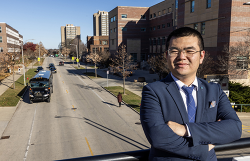 A man with glasses and a navy sportscoat and tie looks at the camera. He is standing on a bridge over traffic.