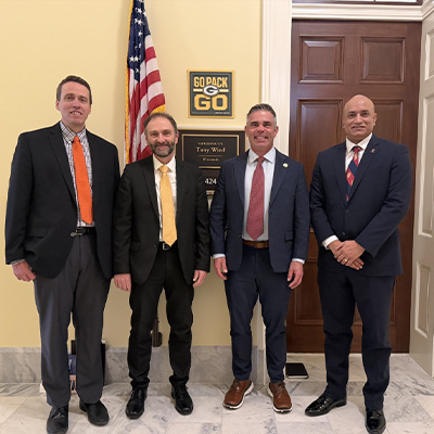 Four men stand in a row looking at the camera. All have on dark suits but different colored ties. One has a beard.