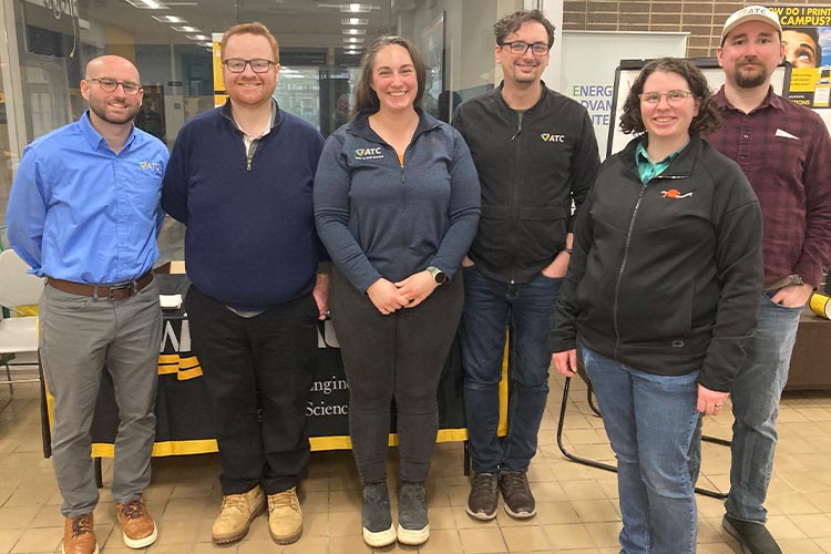 A group shot of six people, including two women. All are wearing dark tops, except for the man on the far left who is wearing a light blue shirt.